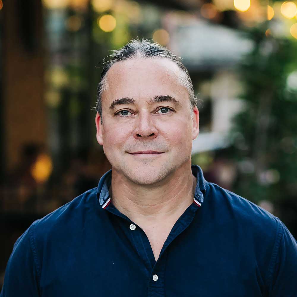 picture of municipal election candidate Jared Mumford in a blue button down dress shirt with sleeves rolled up, and unfocused bokeh lights in the background