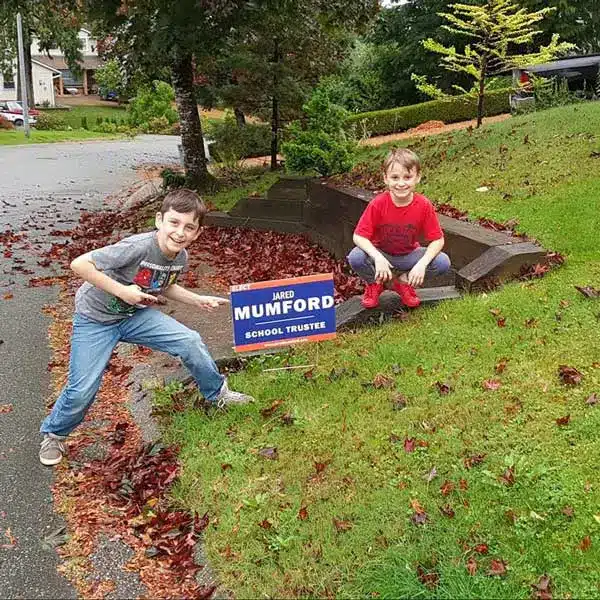 Two boys standing beside, and pointing at an orange and blue sign reading "Vote for Jared Mumord"